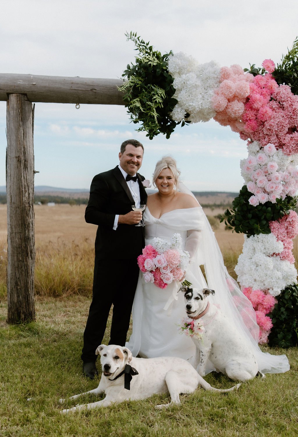 A bride and groom standing under a floral arch on a wedding day, with two dogs at their feet, in a rural outdoor setting.