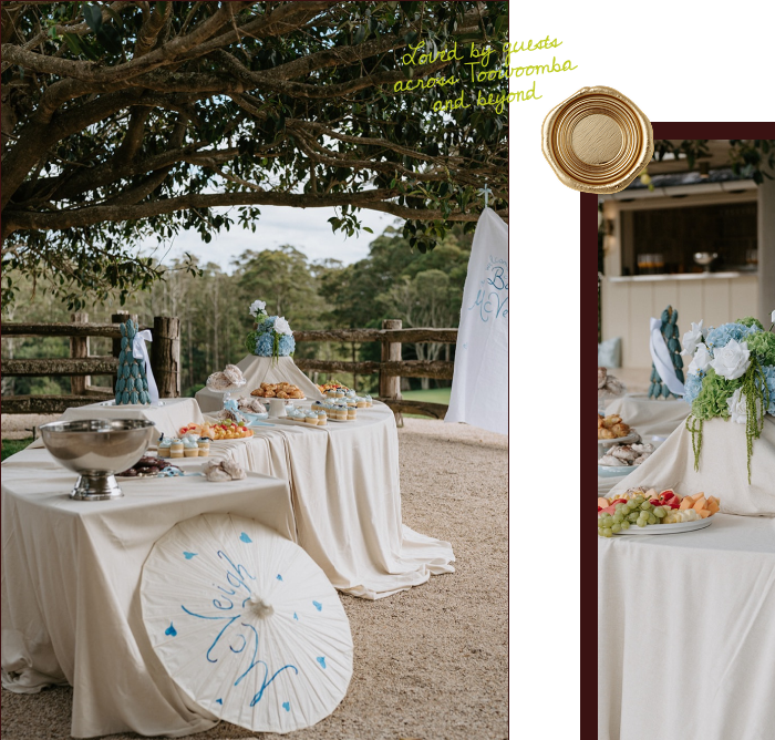 Outdoor dessert table with white tablecloth, flowers, pastries, and fruit, set under a tree with a rustic fence, during daytime.