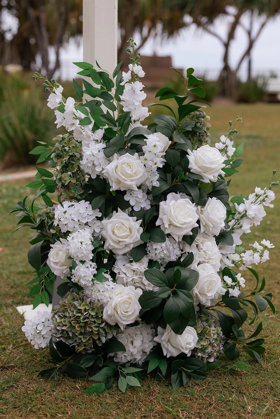 A floral arrangement of white roses, hydrangeas, and other white blossoms with green leaves, outdoors on grass with trees and a cloudy sky in the background.