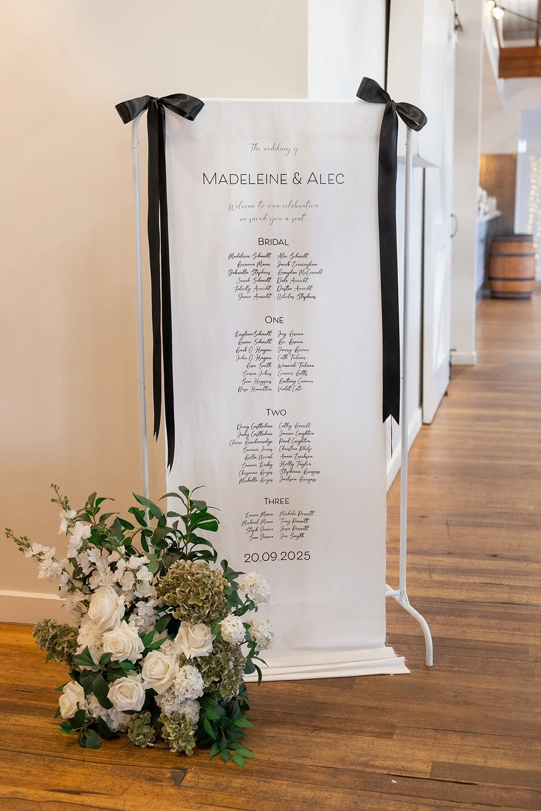 Wedding seating chart with names of guests and a floral arrangement of white roses, hydrangeas, and greenery at the base.