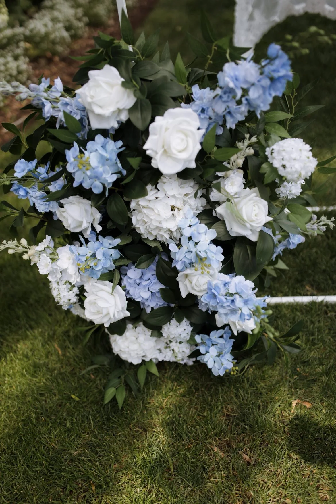 A floral arrangement with white roses, blue hydrangeas, white carnations, and greenery, placed on a grassy surface.