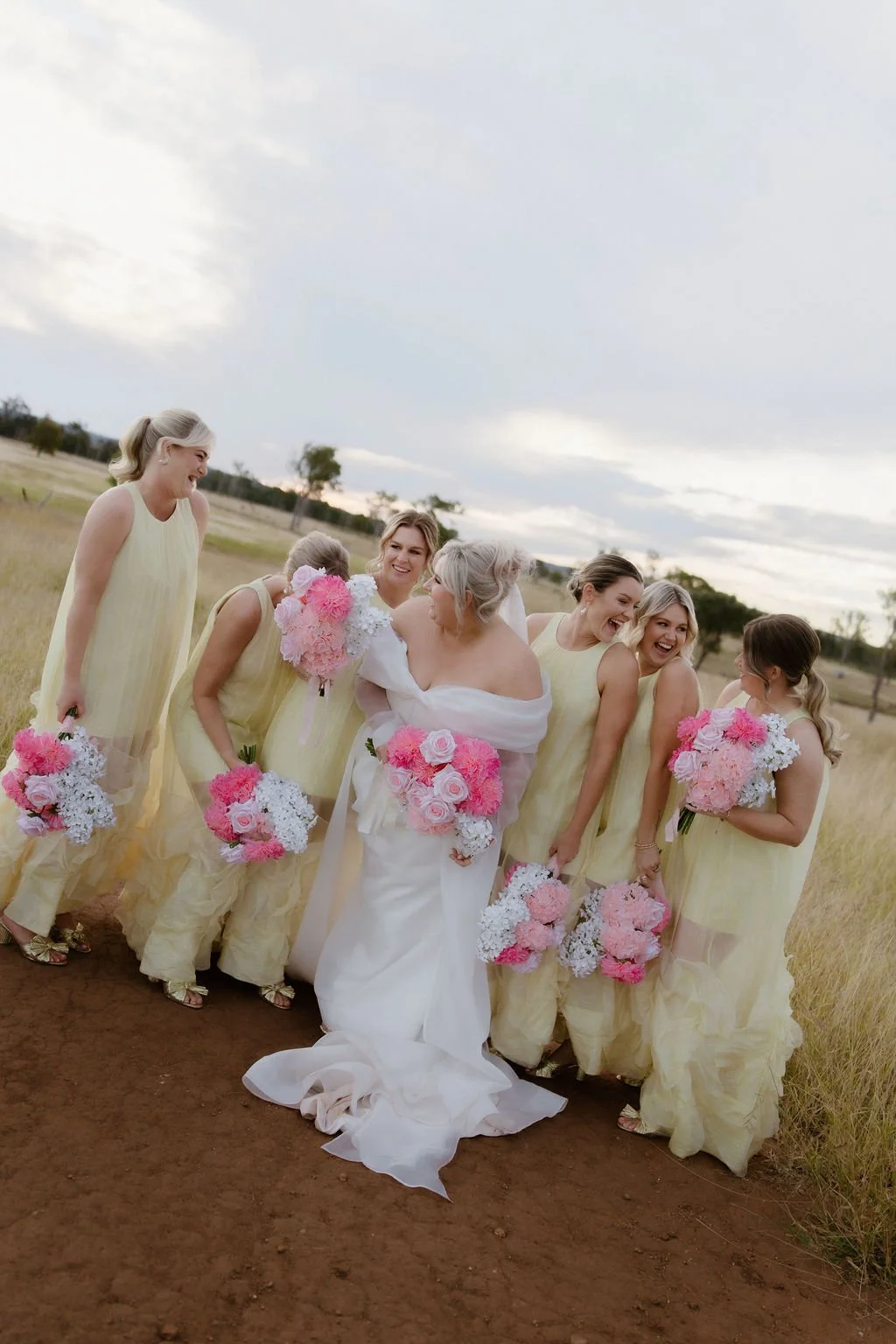 A bride and six bridesmaids in yellow dresses and a bride in a white gown, all holding pink and white bouquets, standing on a dirt path in an open field, smiling and laughing during a wedding photoshoot.