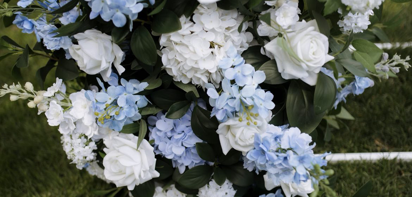 A bouquet of white and light blue flowers, including roses and hydrangeas, with dark green leaves, resting on grass.