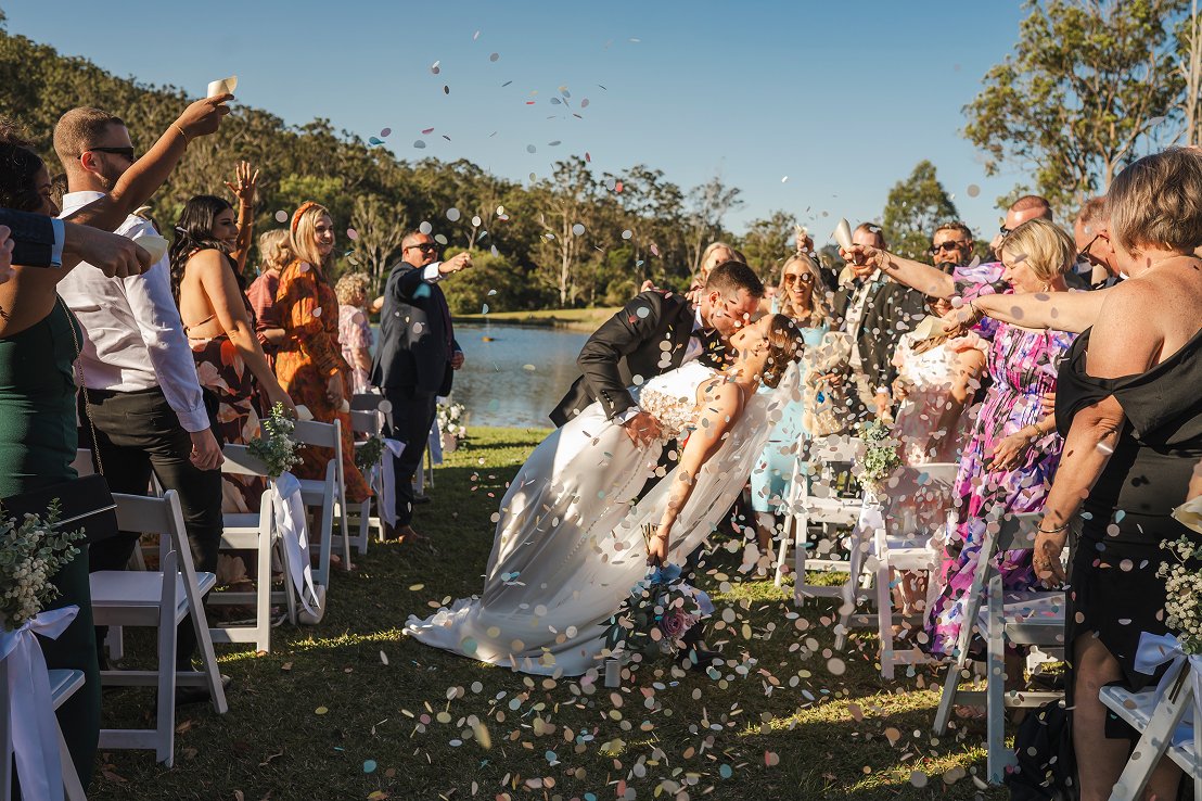Bride and groom kissing under confetti at an outdoor wedding ceremony near a lake, with guests celebrating and throwing confetti.