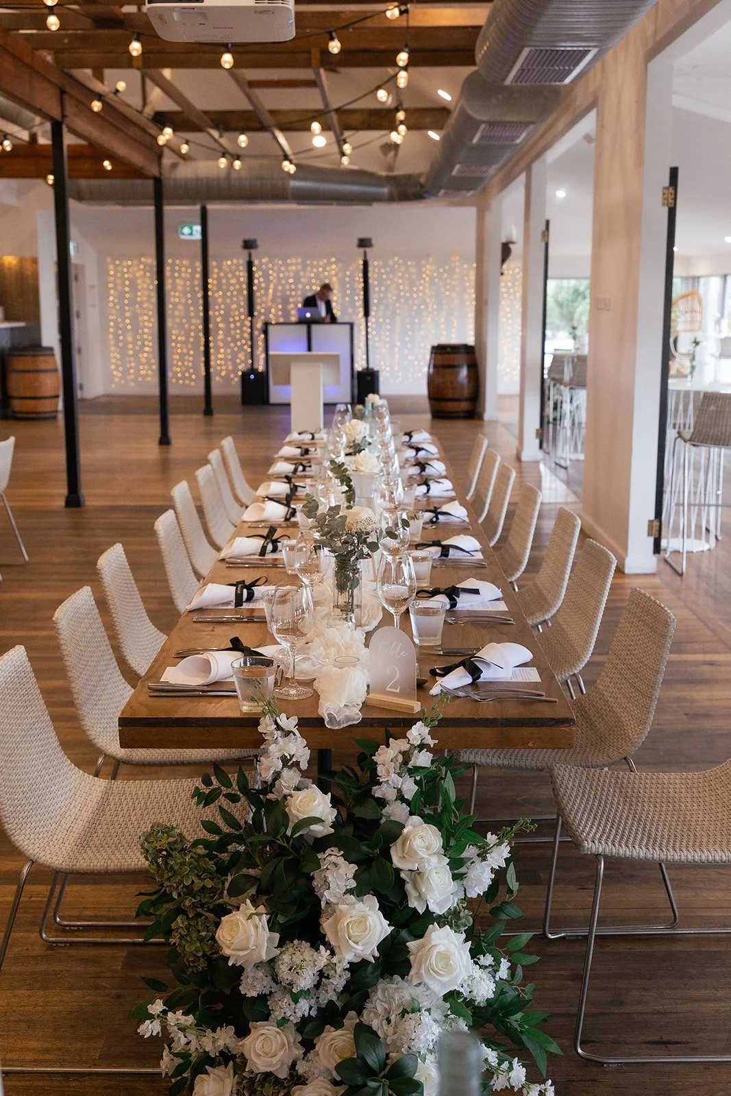 Indoor wedding reception with a long wooden table decorated with white flowers, candles, and glasses, set in a rustic venue with string lights, a DJ booth, and a ceiling with exposed ductwork.