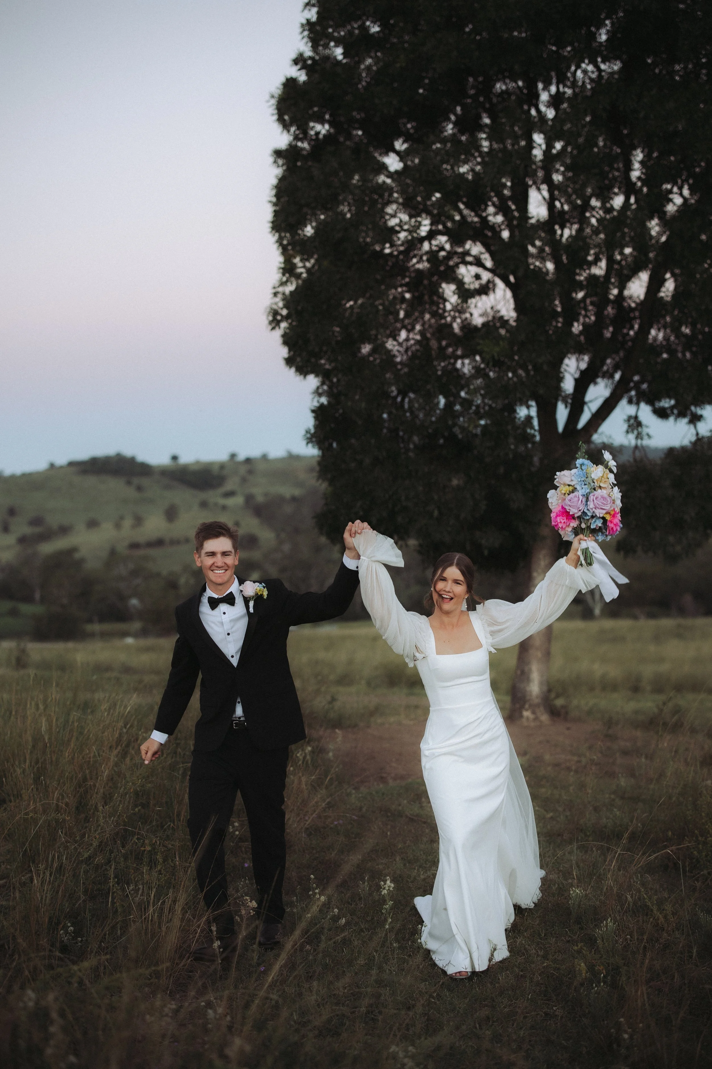 A newlywed couple walking through a grassy field during sunset, holding hands with the bride holding a bouquet of pink, white, and purple flowers. The groom is dressed in a black tuxedo and the bride in a white wedding gown.