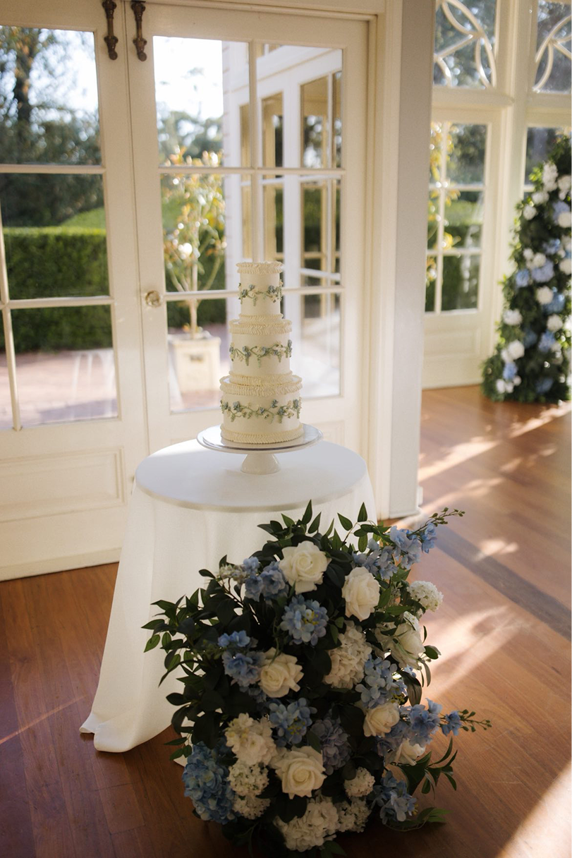 A three-tiered wedding cake decorated with blue and white floral accents on a cake stand, placed on a white round table draped with a white cloth. In front of the table, there is a large bouquet of white and blue flowers with greenery. The setting appears to be indoors near large windows with natural light streaming in.