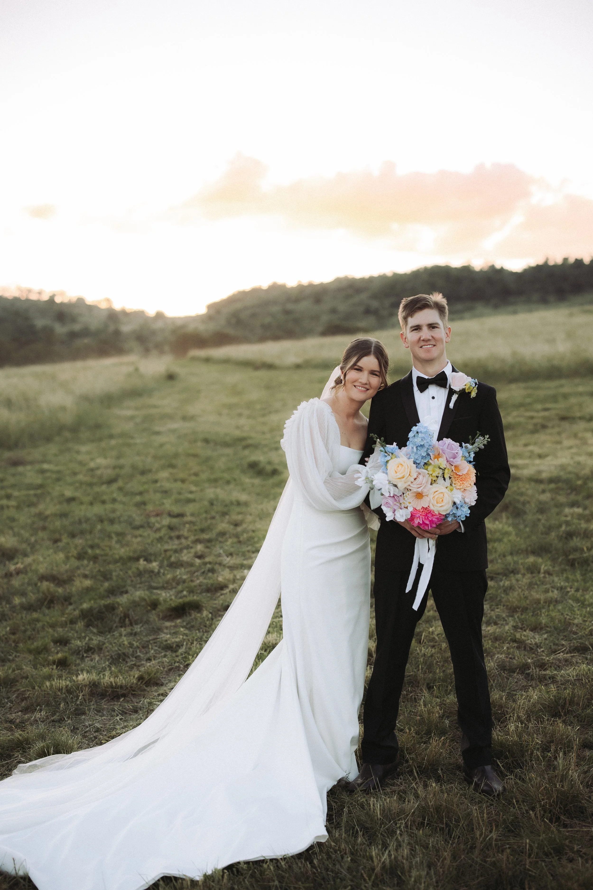 A smiling bride and groom standing in a grassy field at sunset, with the bride holding a bouquet of colorful flowers and wearing a white wedding dress, and the groom in a black tuxedo with a bow tie.