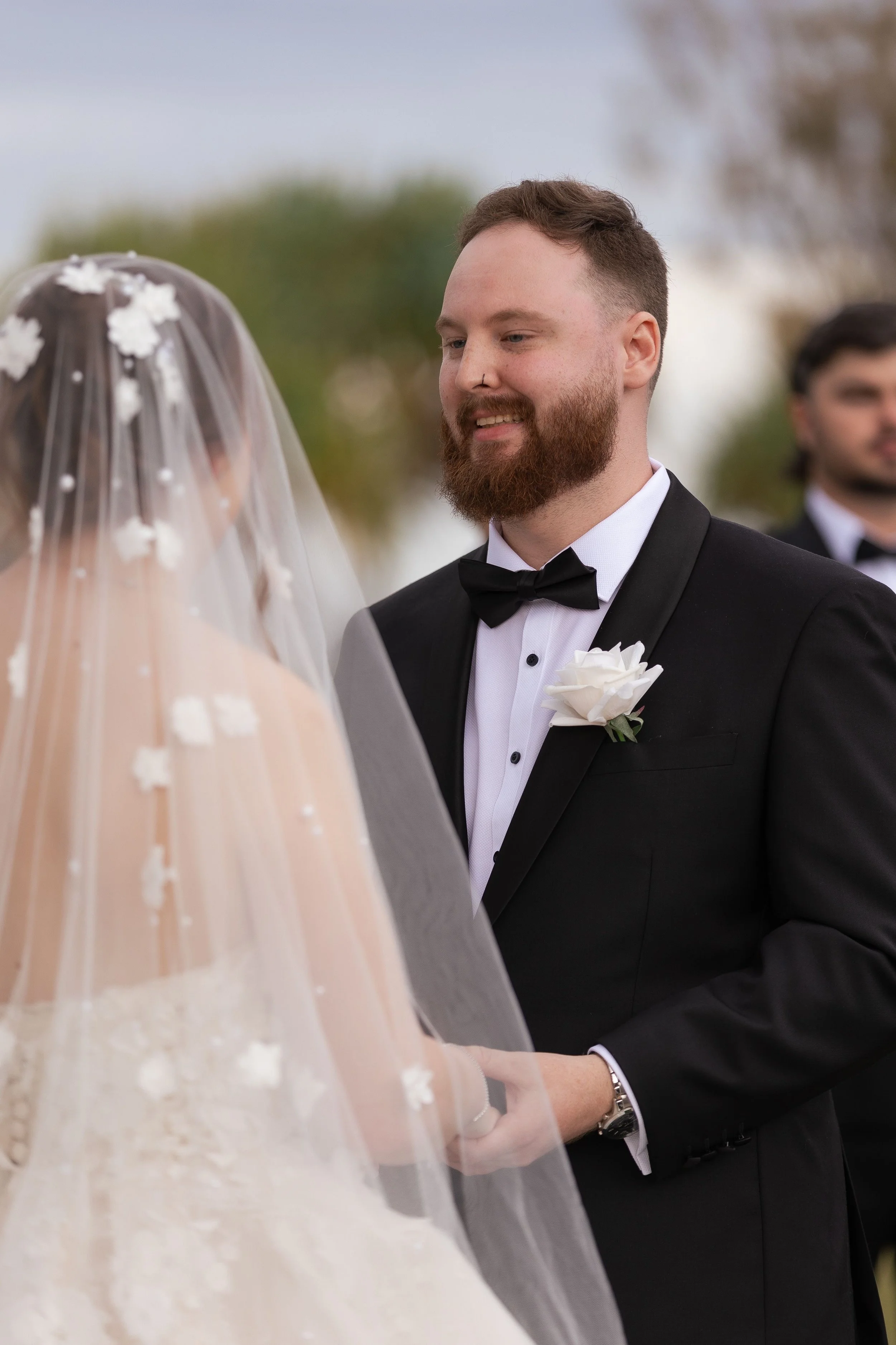 A groom in a black tuxedo with a white boutonniere holds hands with a bride wearing a veil with floral accents during a wedding ceremony outdoors.