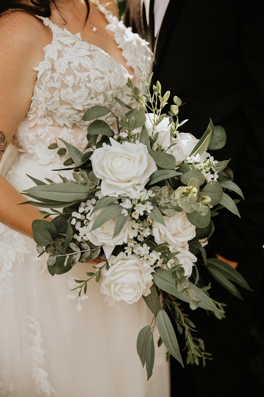 Bride holding a bouquet of white roses and greenery, wearing a lace wedding dress with floral embroidery, and a pearl necklace, standing next to a groom in a black suit.