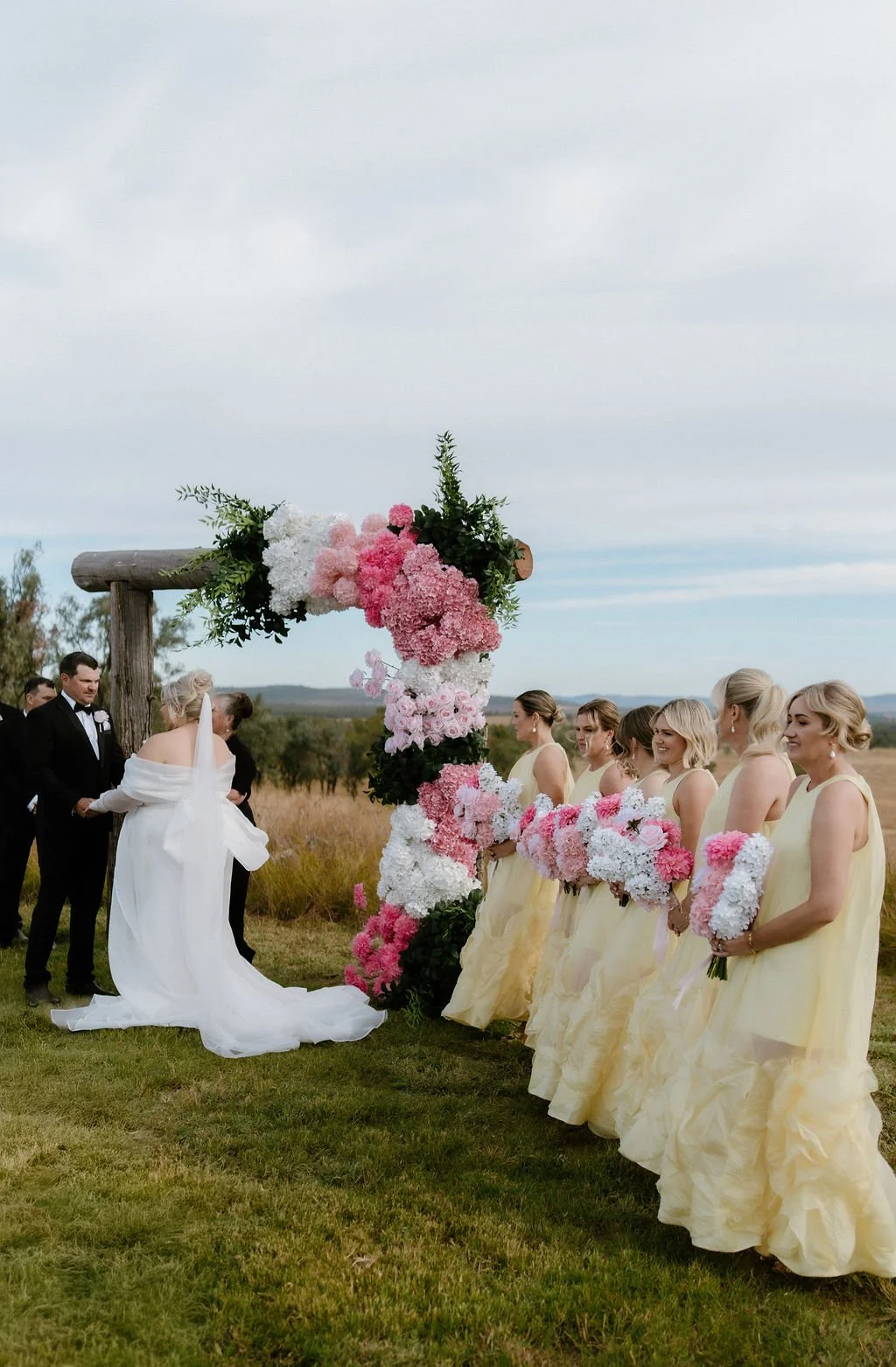 A wedding ceremony outdoors with the bride and groom holding hands under a wooden arch decorated with pink and white flowers. The bride is in a white gown with a veil, and the groom is in a black tuxedo. Bridesmaids in yellow dresses holding bouquets