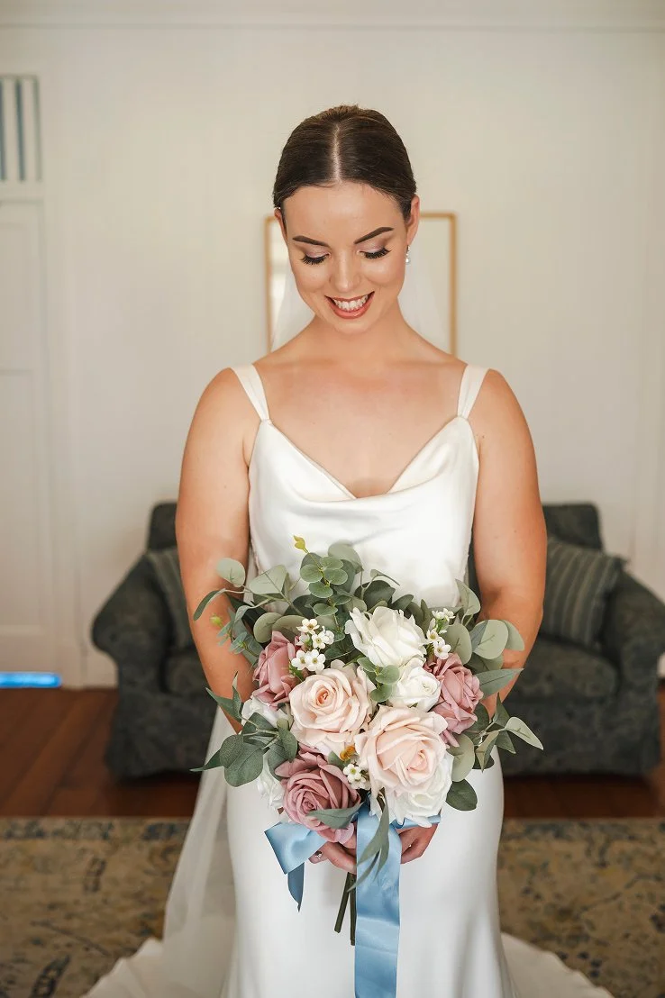 A bride in a white satin dress holding a bouquet of pink and white roses with eucalyptus leaves, standing indoors in front of a couch.