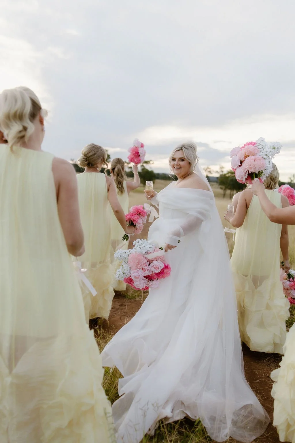 Bride in a white dress holding a bouquet of pink and white flowers, standing among bridesmaids dressed in light yellow, holding pink and white bouquets and flowers, outdoors in a field under cloudy sky.