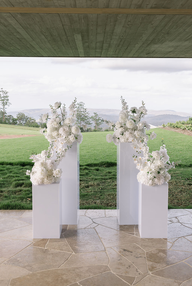 Elegant white floral arrangements on tall white pedestals outdoors under a covered patio, overlooking a green field and distant hills.
