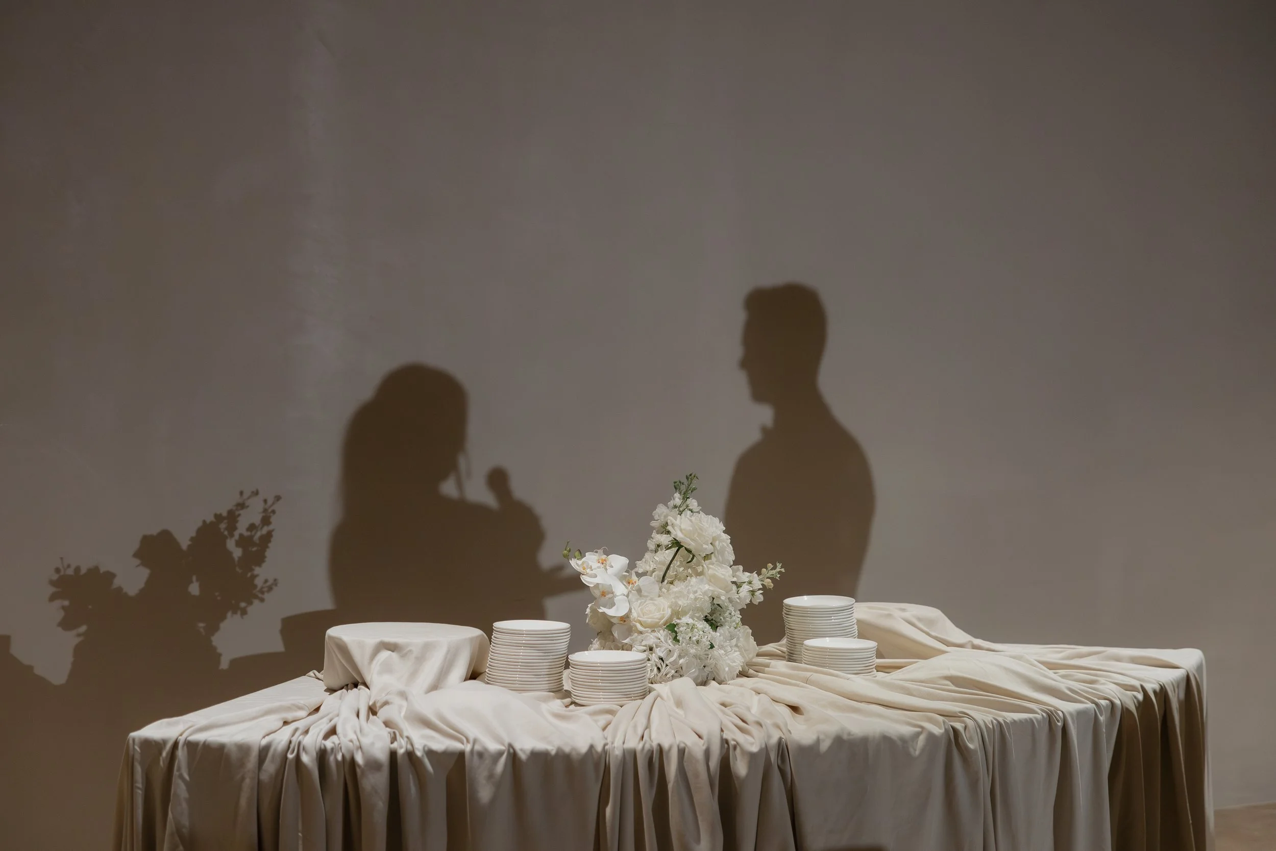 Shadowed silhouette of a man and a woman standing behind a table with white floral arrangements and cream-colored tablecloth, against a plain wall.