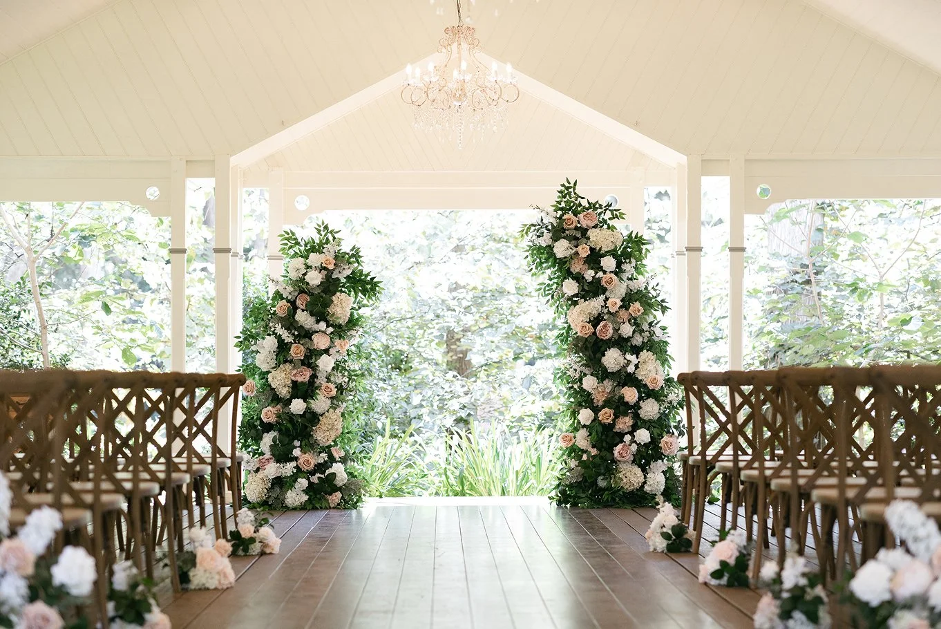 Interior of a wedding ceremony space with floral arches and wooden chairs, overlooking a garden area.