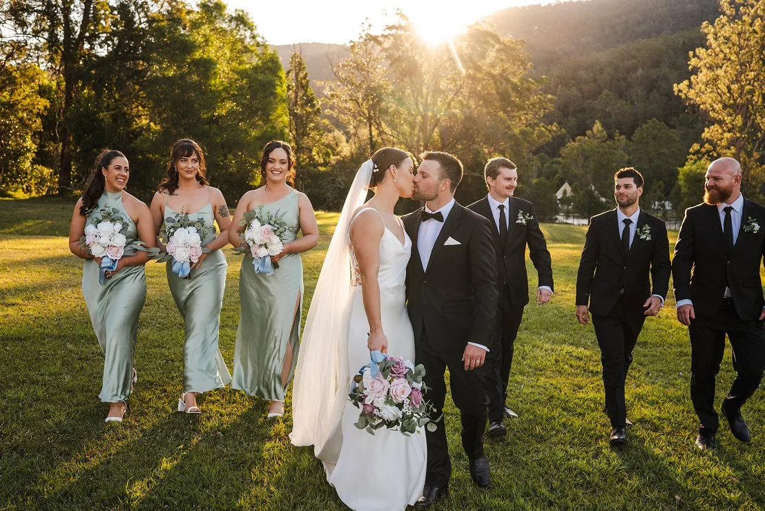 Bride and groom kissing outdoors during sunset, with bridesmaids and groomsmen walking behind them on a grassy field.