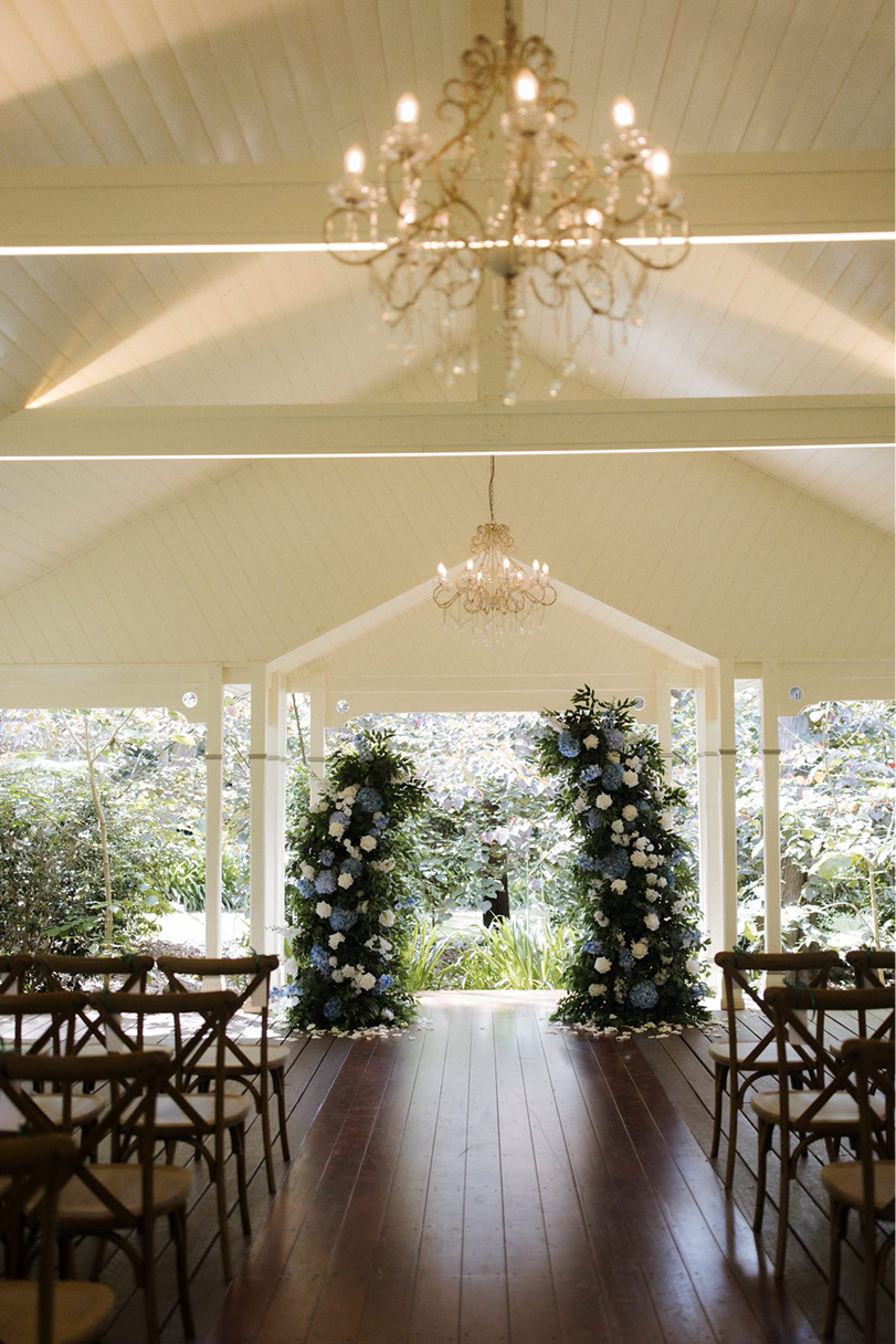 Indoor wedding ceremony setup with rows of chairs facing a floral arch and view of greenery outside, chandeliers hanging from the ceiling.