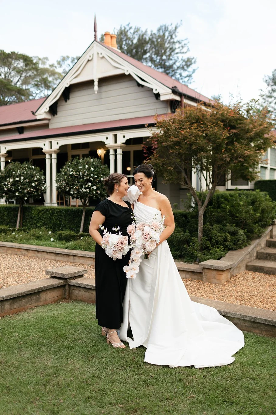Two women, one in a black dress and one in a white wedding gown, smiling and holding bouquets of pink and white flowers, standing on a lawn in front of a large house with a veranda and trees.