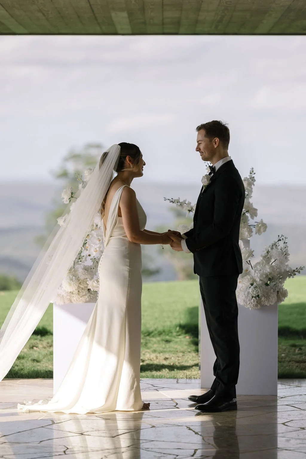 A bride and groom holding hands during their wedding ceremony, standing in front of floral arrangements with a scenic outdoor background.
