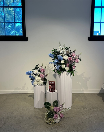 Three white pedestal vases with large floral arrangements of pink, white, and purple flowers and green foliage, placed indoors against a white wall with two windows.