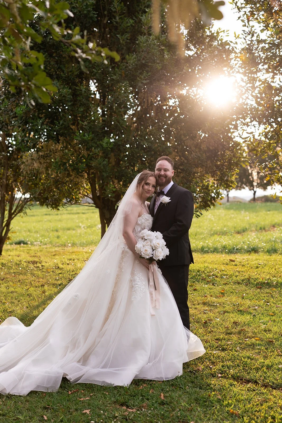 A bride and groom stand together outdoors in a sunlit park, with trees in the background. The bride is in a strapless white wedding gown with a long train, holding a bouquet of white roses. The groom wears a black tuxedo with a white shirt and black 