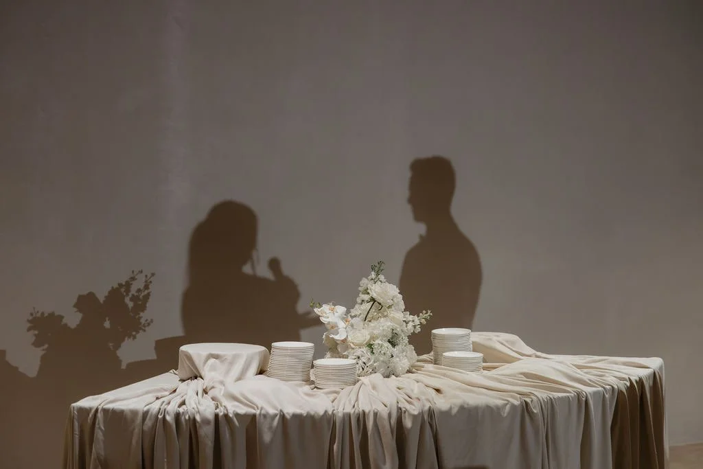 Silhouettes of a woman with a microphone and a man on a plain wall, with a table in front decorated with a white tablecloth, a floral centerpiece, and stacked plates.