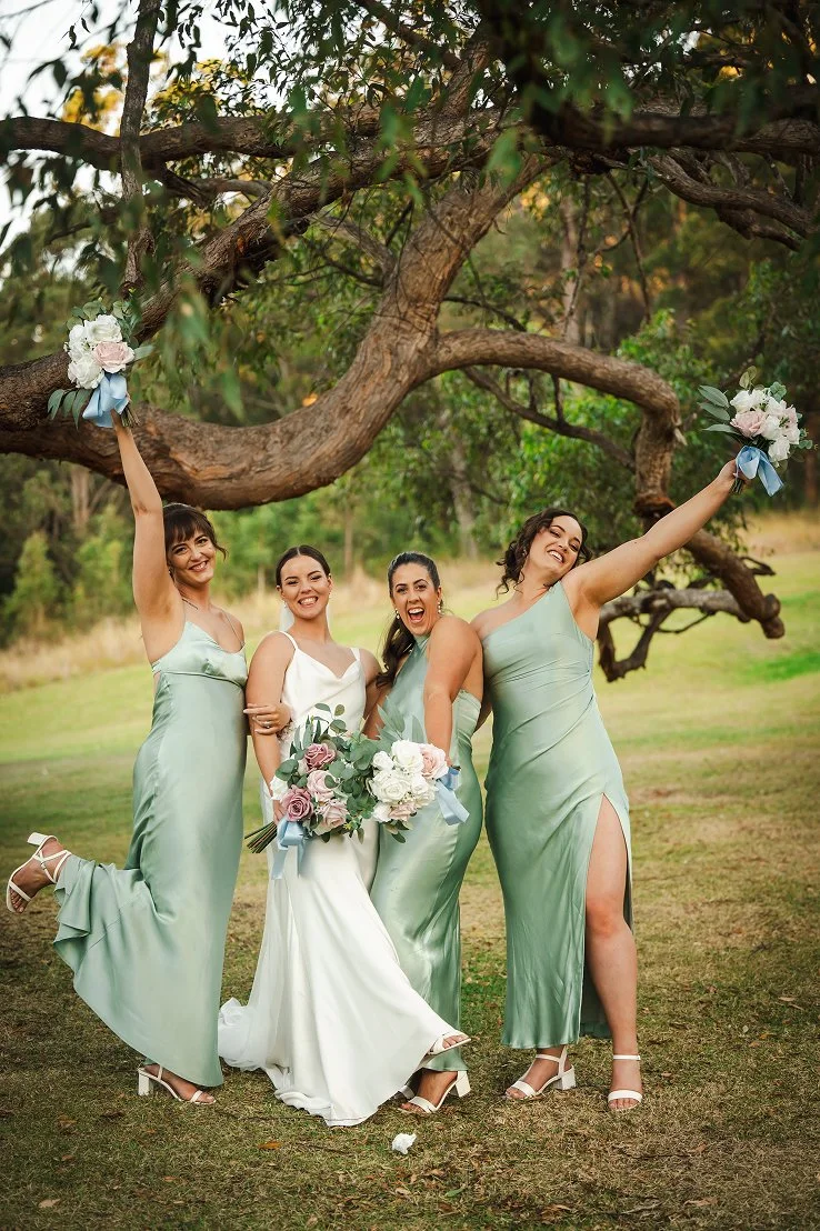 Group of four women in light green and white dresses celebrating outdoors under a large tree, holding bouquets of flowers, smiling and posing happily.