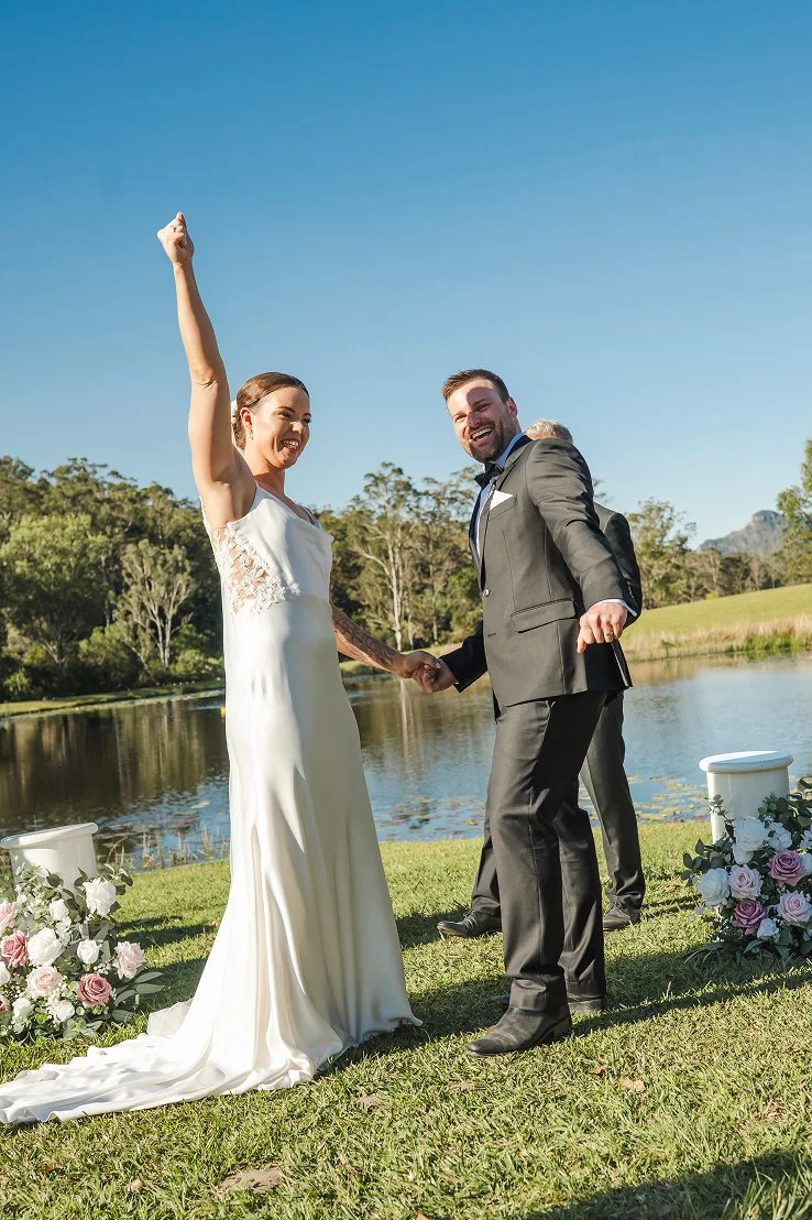 A newly married couple holding hands outdoors near a lake, with trees and mountains in the background, celebrating their wedding.