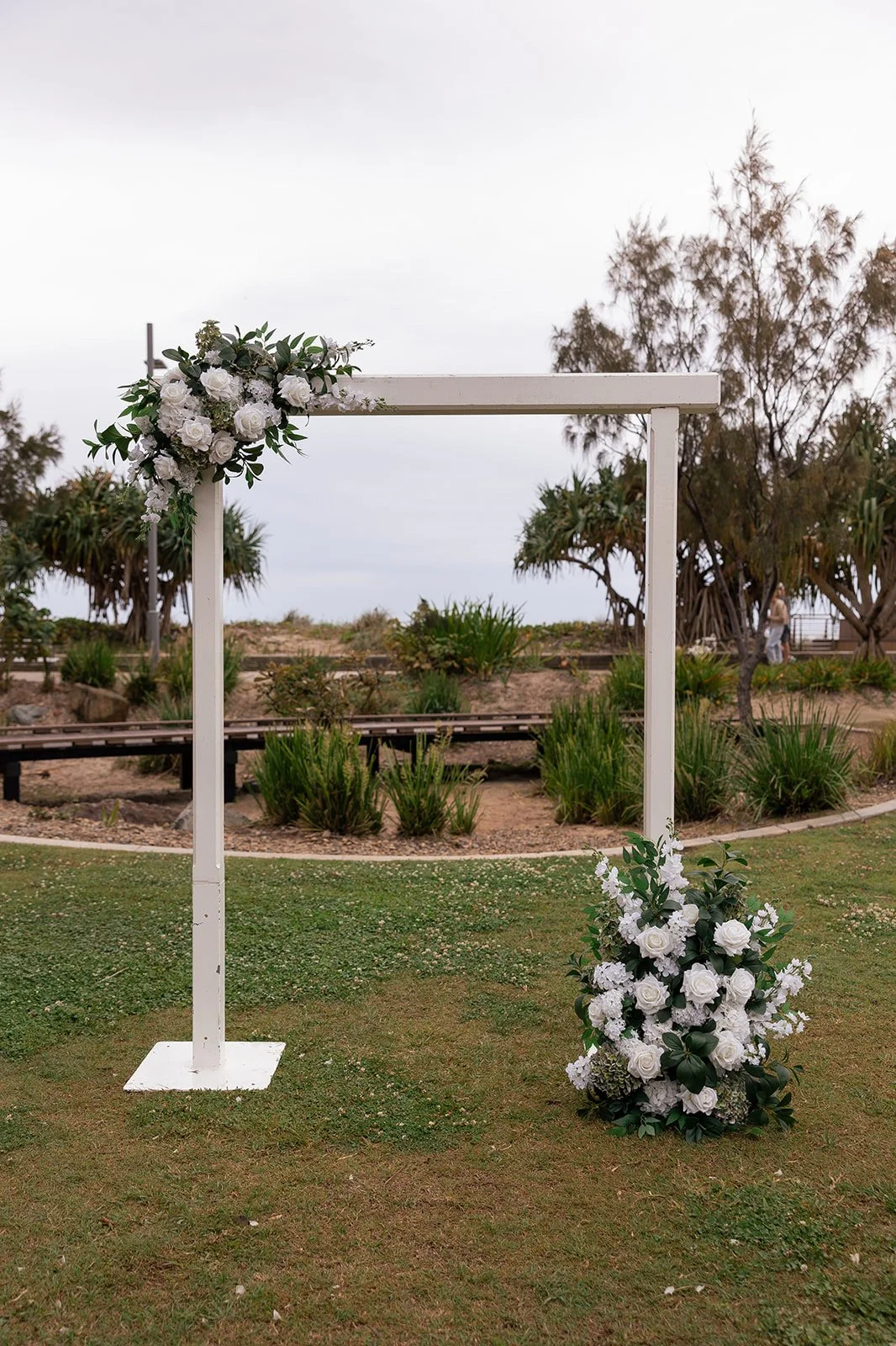 A white wooden wedding arch decorated with white roses and greenery, set outdoors on a grassy area with trees and cloudy sky in the background.
