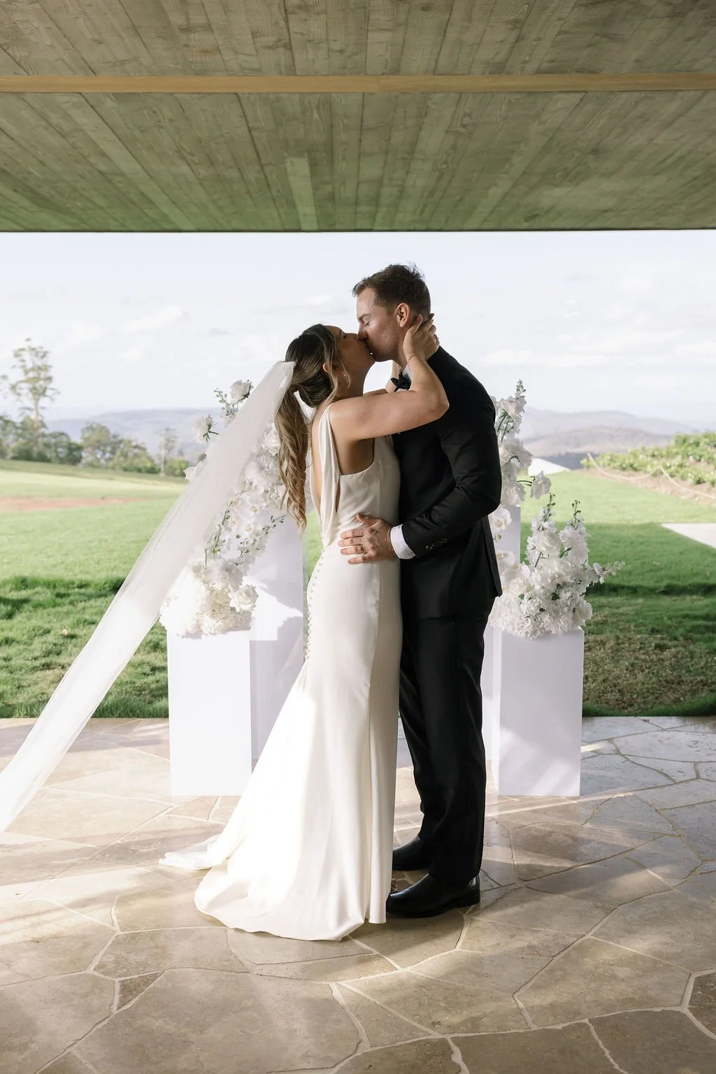 A bride and groom share a kiss at their wedding ceremony outdoors, with white floral decorations behind them and green fields in the background.