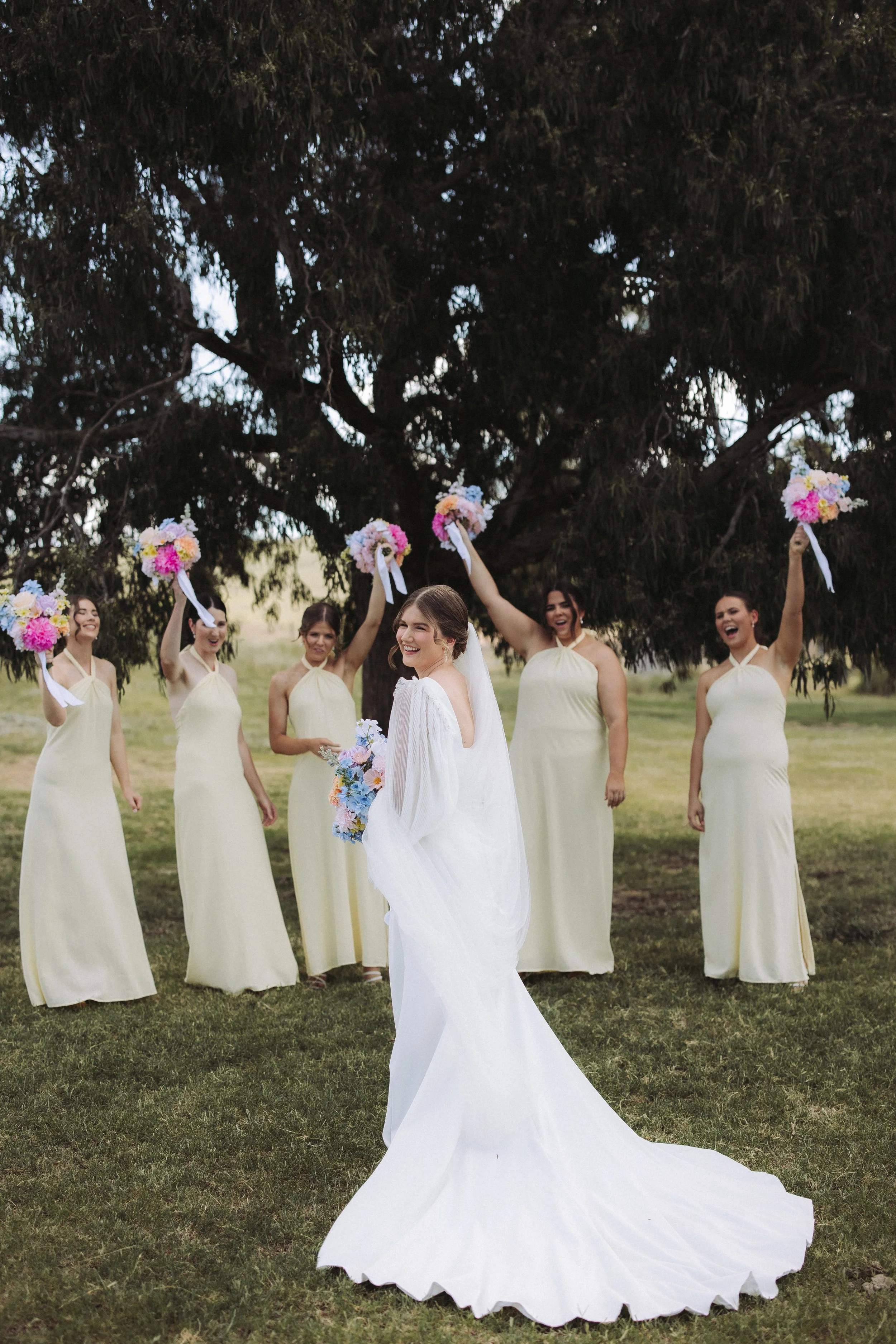 Bride in white wedding dress smiling with five bridesmaids in matching yellow dresses holding bouquet flowers, outdoors under a large tree.