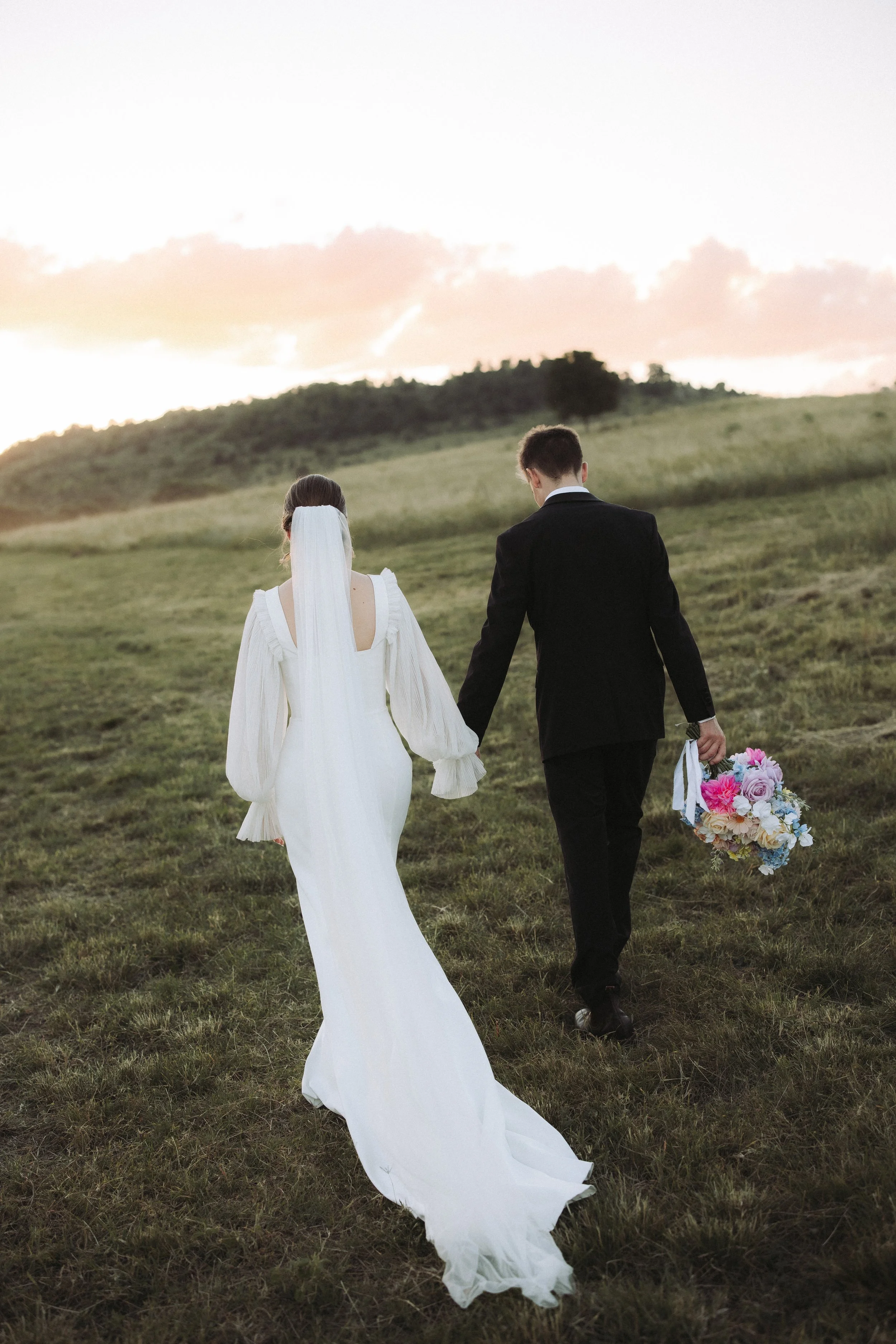 A bride and groom walking hand in hand across a grassy field at sunset, with the bride holding a bouquet of pink and purple flowers.