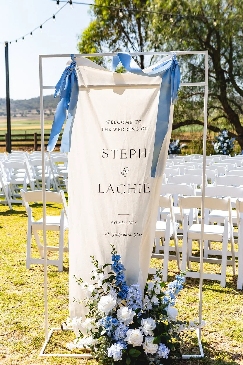 Wedding welcome sign with white fabric, blue ribbons, and floral arrangement at an outdoor ceremony with white chairs and string lights