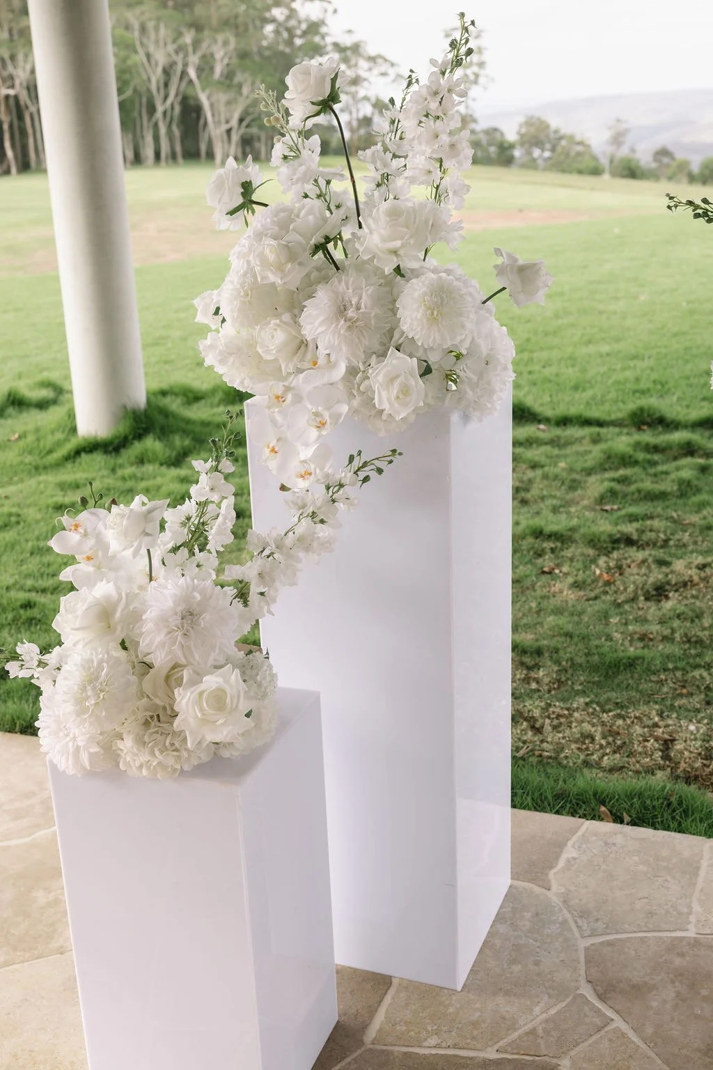 White floral arrangements on two white pedestals outdoors with a grassy field and trees in the background.