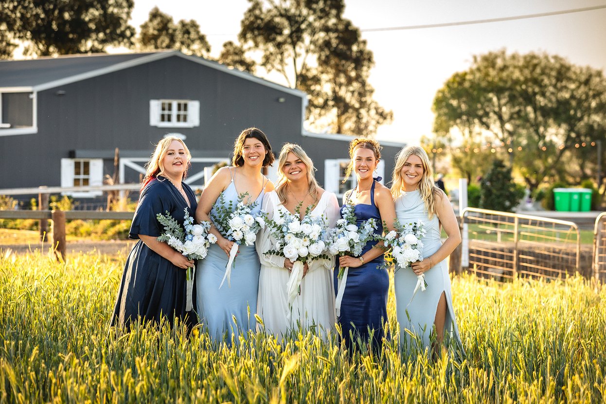 Five women dressed in formal dresses, holding bouquets of white and green flowers, standing in a grassy field with a barn and trees in the background during sunset.