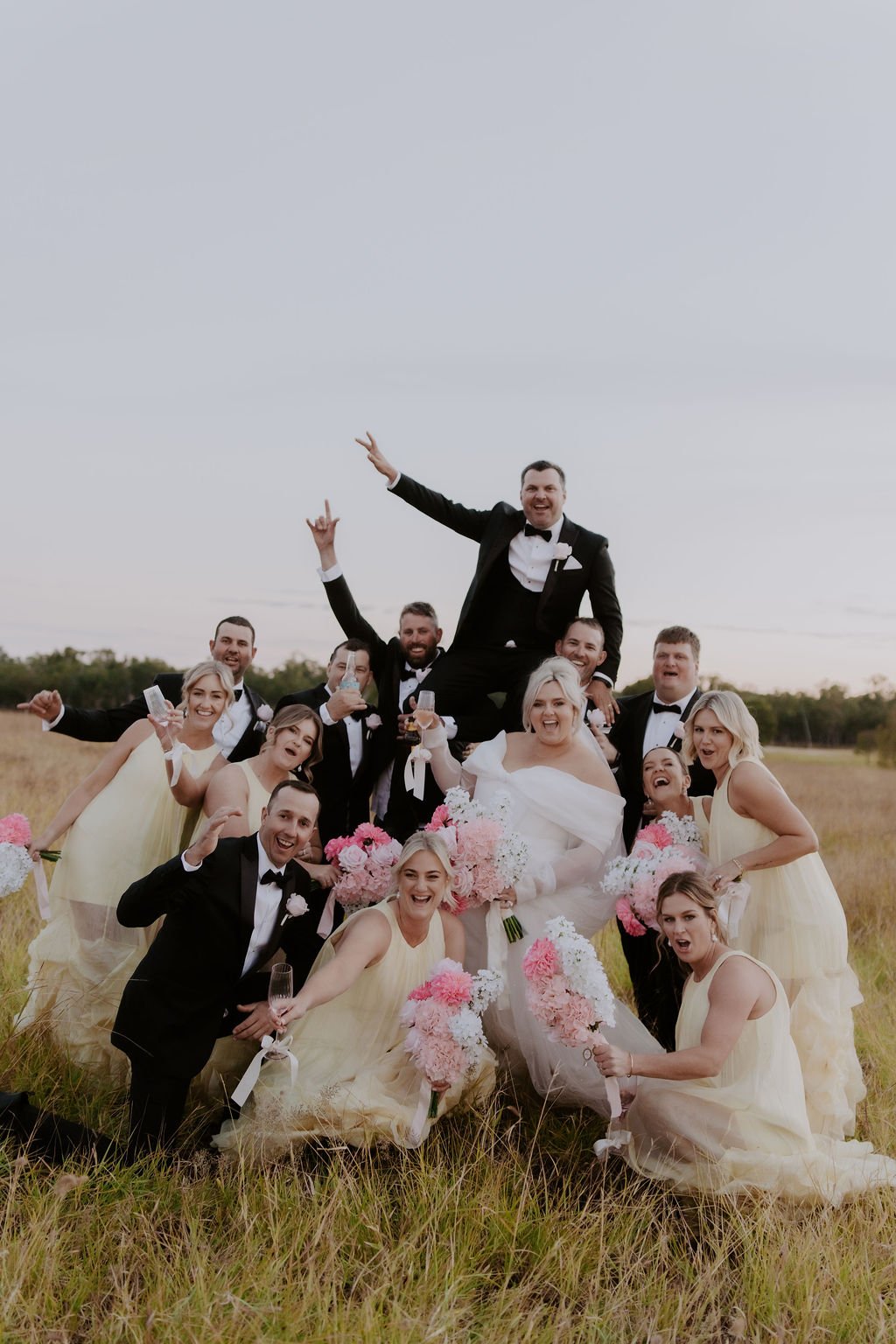 A wedding celebration photo in a field with a bride, groom, bridesmaids, and groomsmen, some lifting the groom in a tuxedo, with bouquets of pink and white flowers, all smiling and cheering.