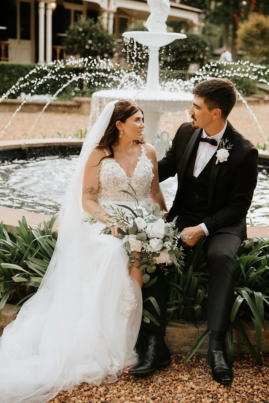 A bride and groom sitting together in front of a fountain during their wedding, with the bride holding a bouquet and wearing a white lace wedding gown, and the groom in a black tuxedo with a white shirt and black bow tie.
