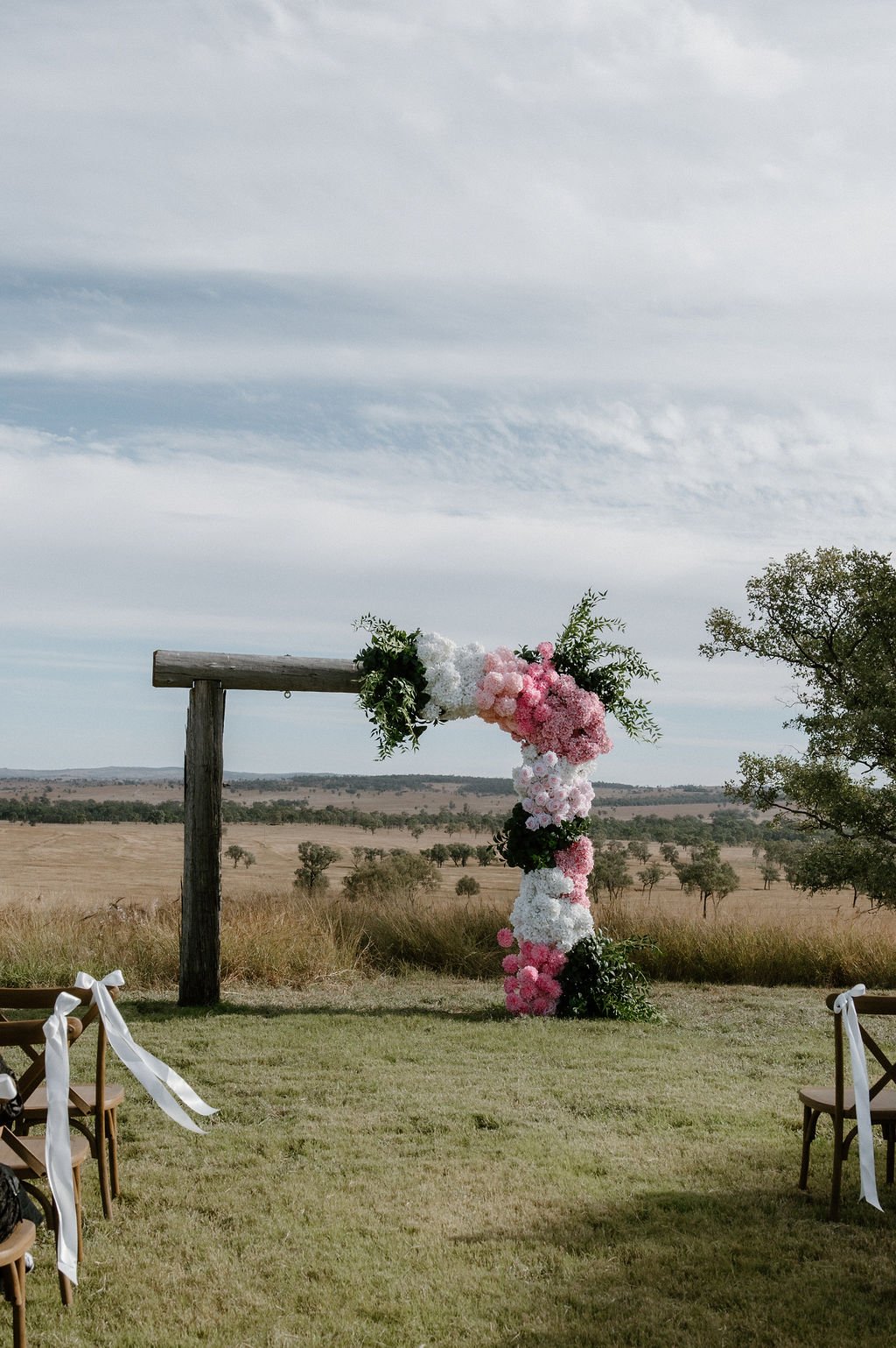 Wedding arch decorated with pink and white flowers and greenery, positioned outdoors on a grassy field with chairs on either side, a tree to the right, and open plains under a cloudy sky in the background.