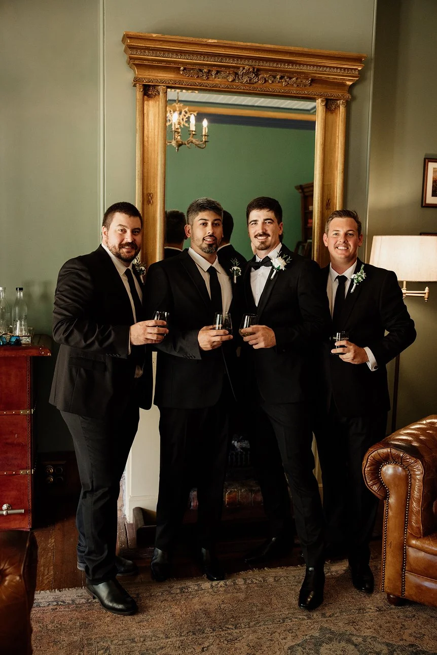 Four men in tuxedos standing together with drinks in hand, smiling in front of a large mirror in an upscale room with vintage decor and a chandelier.