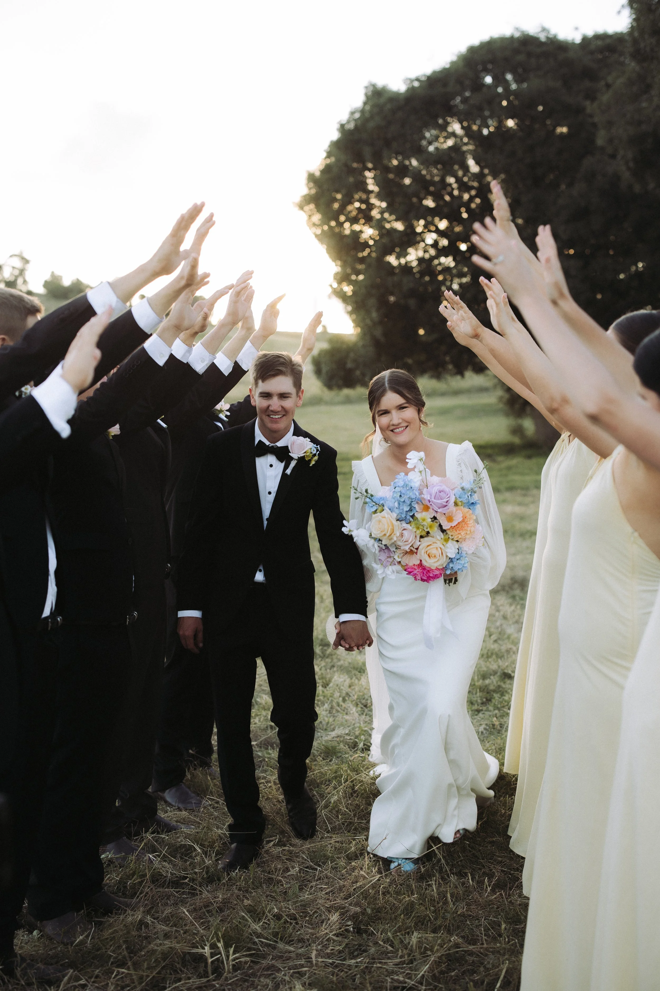 A bride and groom holding hands walking outdoors surrounded by wedding guests with their arms raised, during sunset in a grassy field with trees in the background.