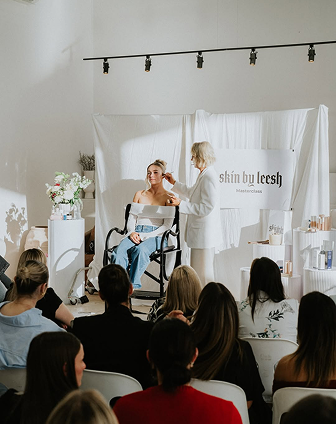 A woman is seated in a wheelchair on stage at a skincare event, speaking into a microphone. An older woman, possibly the speaker or presenter, is standing next to her. Audience members are seated in front, watching the presentation in a room decorated with a skincare banner and product displays.