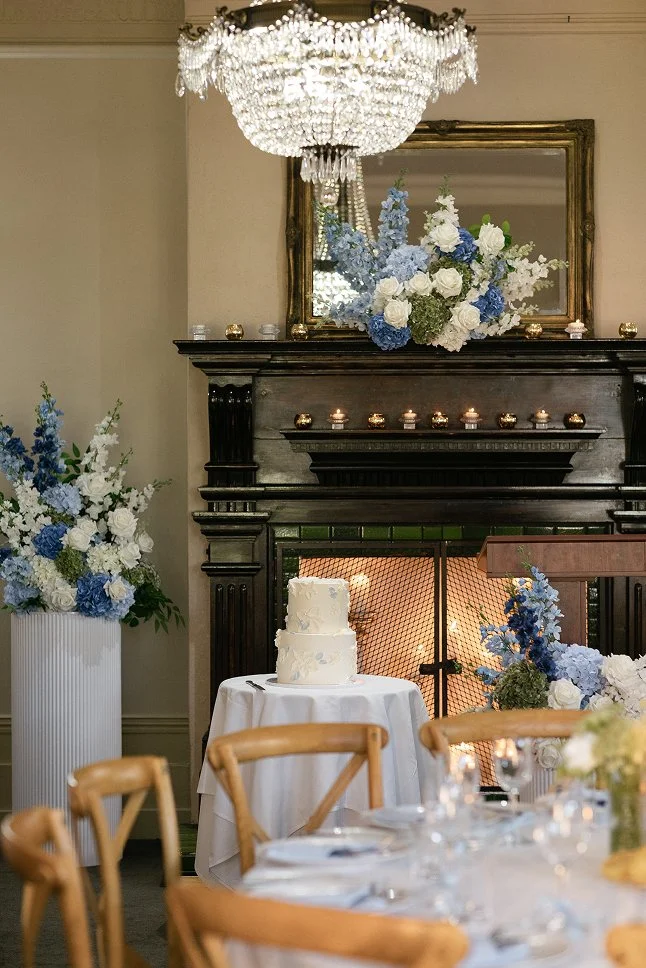 Elegant room decorated for a celebration with a white wedding cake on a small round table, large flower arrangements in white and blue around a fireplace, a crystal chandelier overhead, and a large mirror reflecting the floral display.