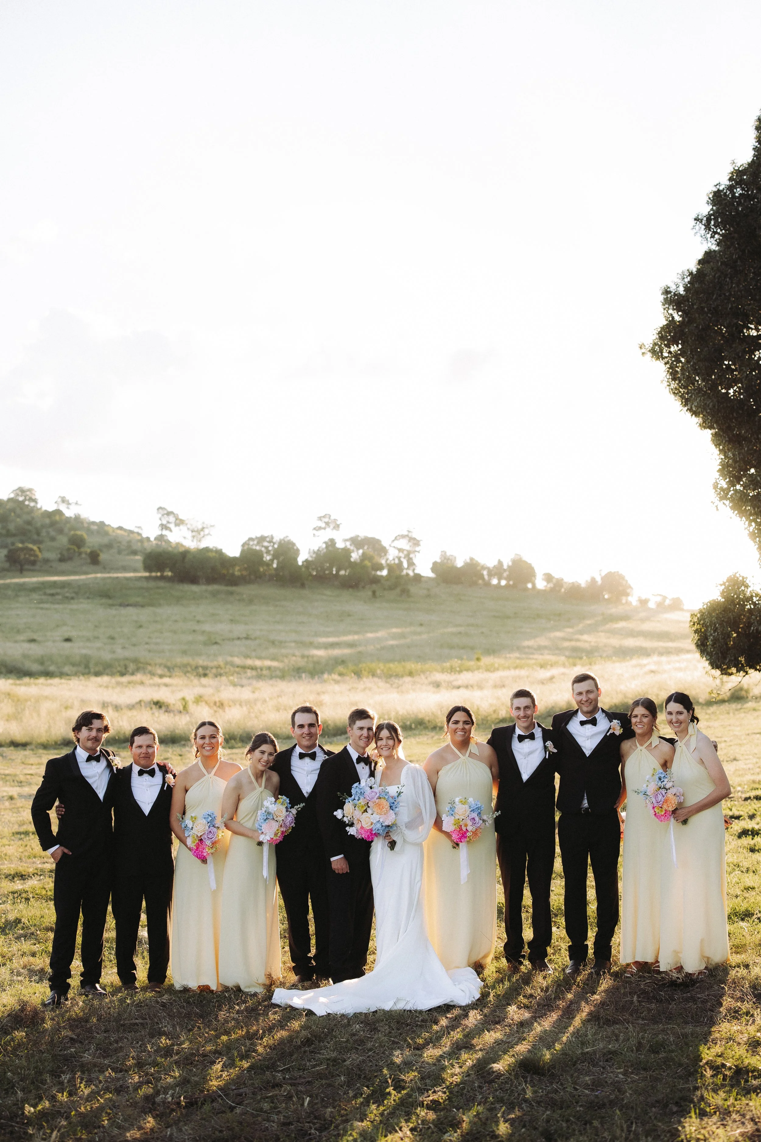 Wedding party consisting of eleven people, including bride and groom, posing outdoors on a field during sunset with hills and trees in the background.