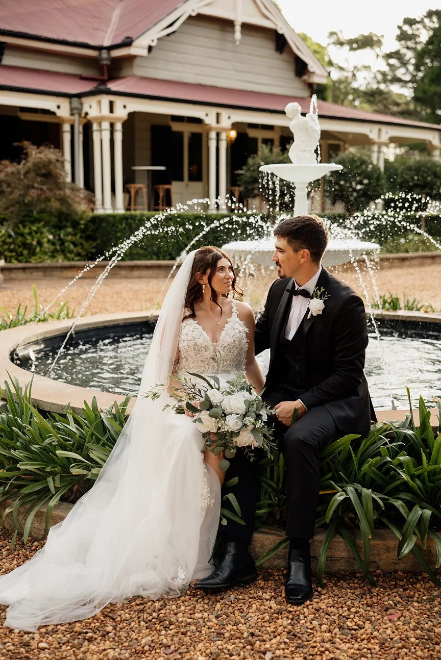 Bride and groom sitting in front of a fountain at a wedding, with bride holding a bouquet of white flowers, and both are dressed in formal wedding attire.