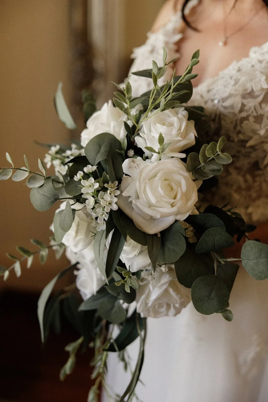 Close-up of a woman in a wedding dress holding a bouquet of white roses, greenery, and small white flowers.