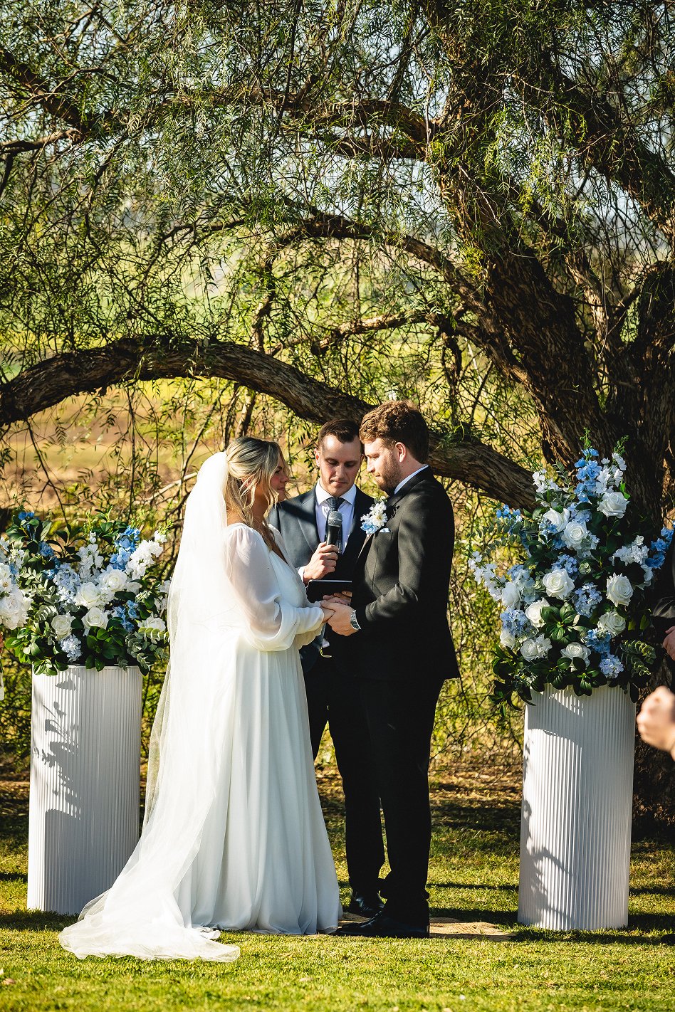 A wedding ceremony taking place outdoors under a large tree with green leaves. The bride and groom are facing each other, holding hands, with a person officiating them speech with a microphone. The bride is in a white wedding dress with a veil, and t