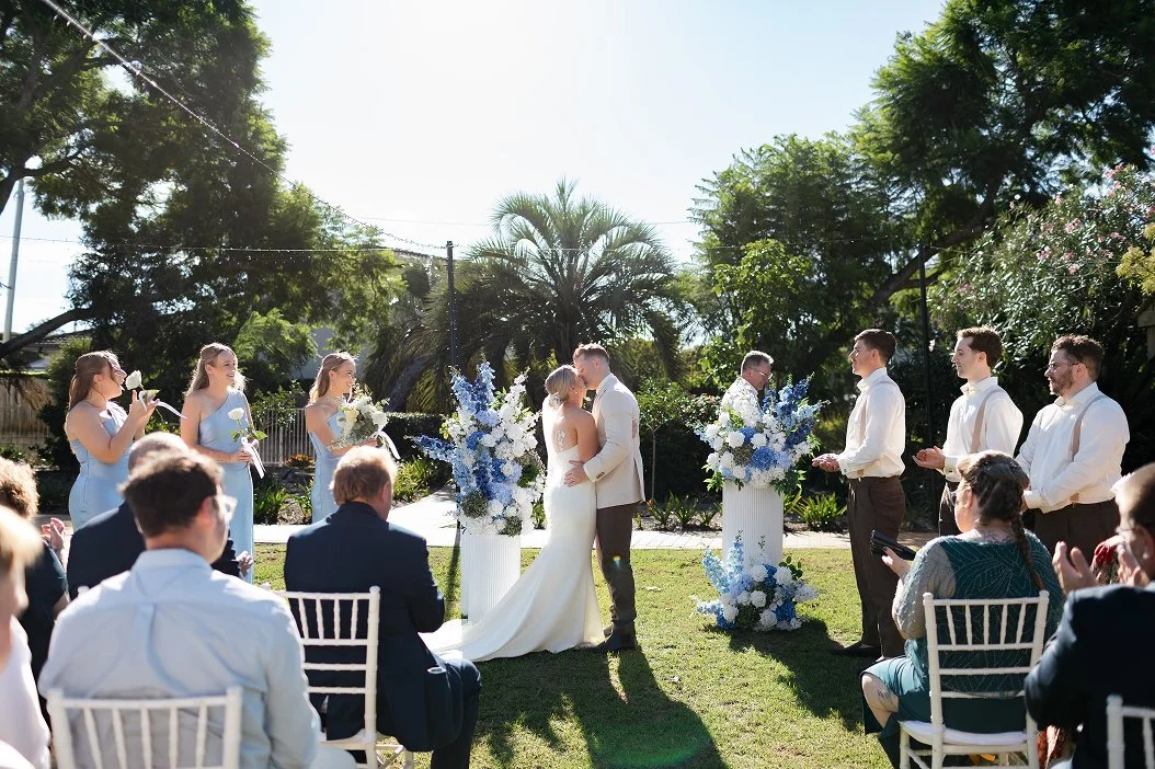 Outdoor wedding ceremony with bride and groom kissing, surrounded by bridesmaids and groomsmen, in a garden with trees and blue skies.