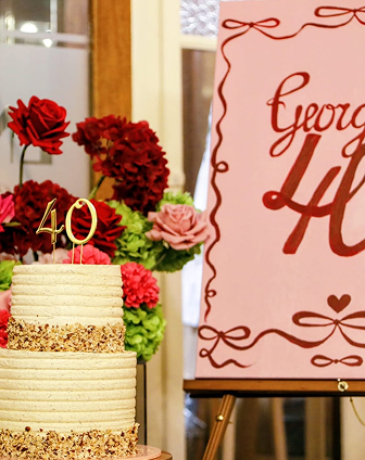 A birthday cake with a 40 candle, decorated with cream and crumb layers, is placed next to a floral arrangement with red, pink, and green flowers, and a sign that says 'George Liv.'
