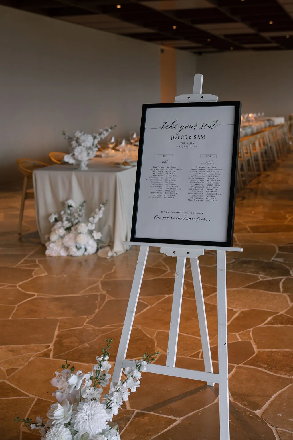 Wedding seating chart on a stand with floral decoration in a reception hall, with a decorated table and chairs in the background.